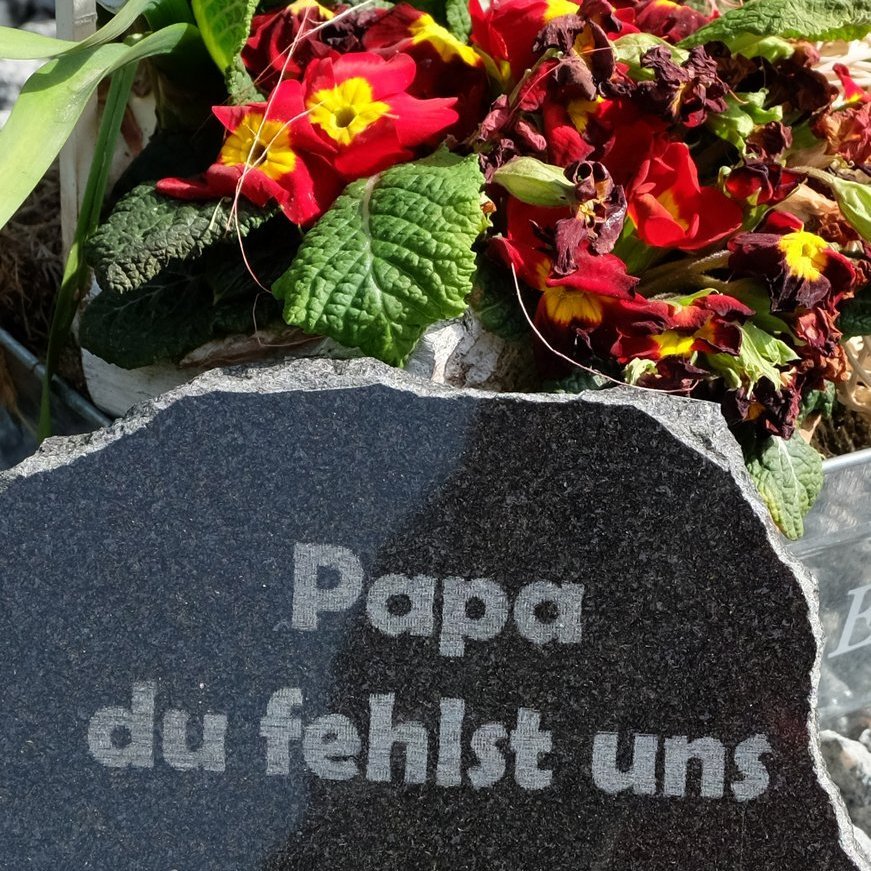 Grabstein vor Blumen mit Aufschrift zum Gedenken an den verstorbenen Vater